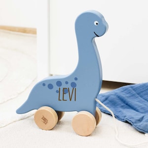 Op de afbeelding: Een blauw houten trek speelgoed in de vorm van een dinosaurus met de naam "Levi" erop geschreven. Het speelgoed heeft bruine houten wielen en een wit trekkoord.
