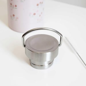May include: A stainless steel water bottle lid with a silver handle. The lid is round and has a brushed metal finish. A pink water bottle with a floral pattern is visible in the background. A cleaning brush is also visible.