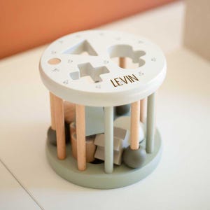 Könnte beinhalten: Ein Holz-Formensortierspielzeug mit hellgrüner Basis und Oberseite, mit ausgeschnittenen Formen und dem Namen "LEVIN". Das Spielzeug hat Holzdübel und enthält verschiedene geometrische Formen im Inneren. Das Design ist einfach und kindgerecht.
