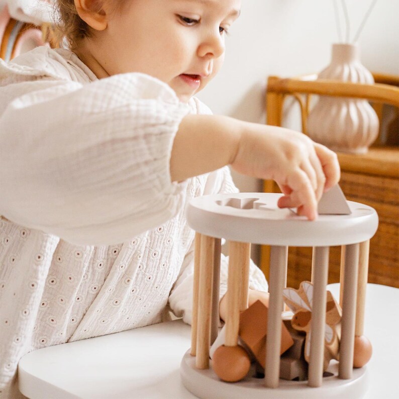 May include: A child plays with a wooden toy with a variety of shapes and colors. The toy has a round base and a cylindrical cage with wooden dowels. The child is holding a wooden shape in their hand and is about to place it into the cage.