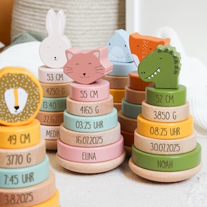 Op de afbeelding: Houten stapelspeelgoed met dierenfiguren, waaronder een leeuw, konijn, kat, haai, vos en dinosaurus. Elke stapel heeft gekleurde ringen met afmetingen in centimeters, grammen en data. De naam "ELINA" staat erop.