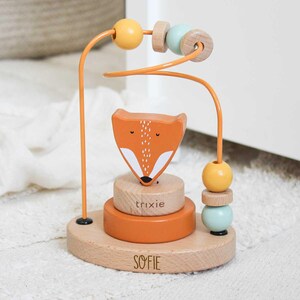 Może przedstawiać: Drewniana zabawka z labiryntem z koralików z pomarańczową głową lisa, żółtymi i niebieskimi koralikami oraz napisami "SOFIE" i "trixie". Zabawka ma pomarańczową drucianą ramę i jest osadzona na drewnianej podstawie. Dla dzieci.