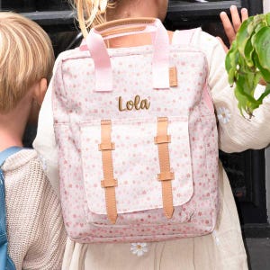 May include: A pink floral backpack with the name "Lola" embroidered in gold. The backpack has light pink straps and accents, with tan leather-like buckles and handle. The bag is designed with a front pocket and a top handle.