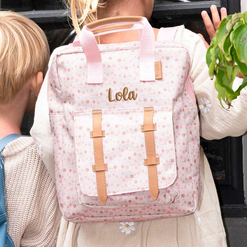 May include: A pink floral backpack with the name "Lola" embroidered in gold. The backpack has light pink straps and accents, with tan leather-like buckles and handle. The bag is designed with a front pocket and a top handle.