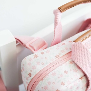 May include: A close-up of a pink and white floral patterned bag with a matching pink strap and a brown leather handle. The bag has a zipper closure and a pink strap with a plastic buckle. The bag is likely a tote or travel bag.