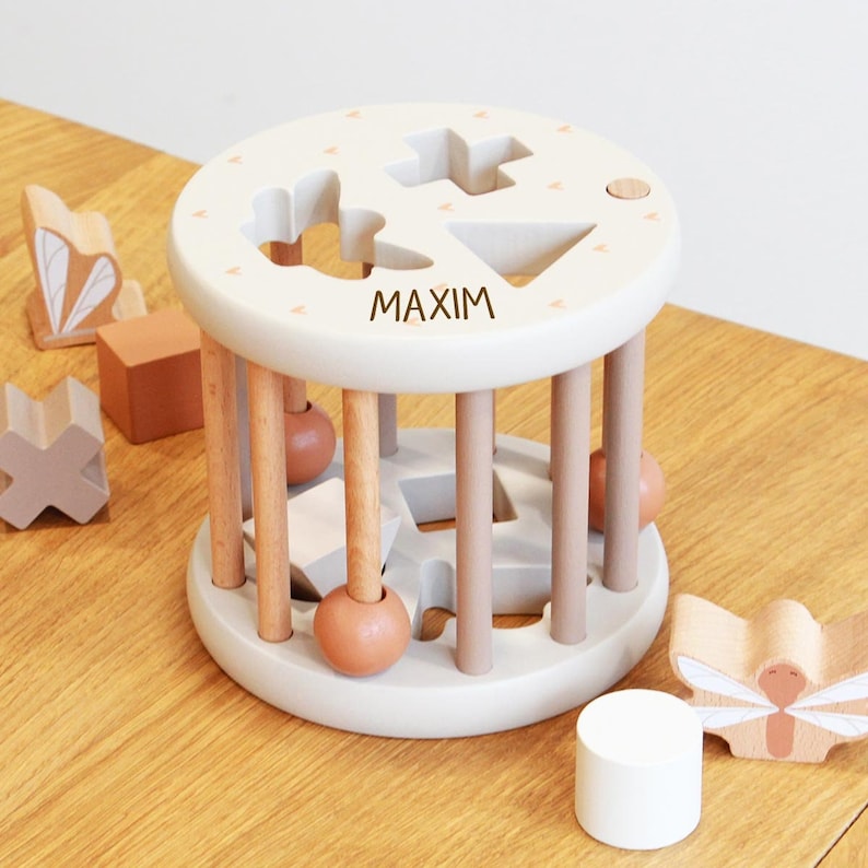 May include: A wooden toy with a rolling cylinder and a removable top. The top has a variety of shapes cut out and the cylinder has wooden dowels that allow for the shapes to be rolled around. The toy is personalized with the name "MAXIM".