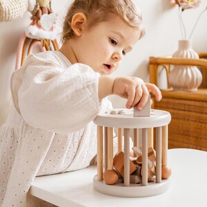 May include: A child plays with a wooden toy with a beige finish. The toy has a cylindrical shape with wooden dowels and a variety of wooden shapes inside. The child is holding a wooden block in their hand.