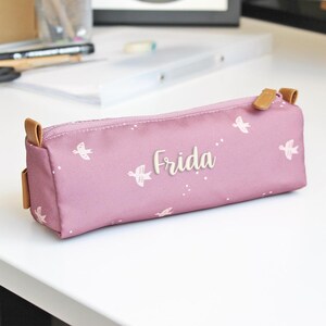 Peut inclure: Une trousse à crayons violette avec un motif d'oiseaux blancs et le nom "Frida" brodé sur le devant.