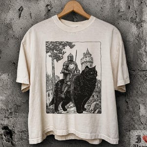 Könnte beinhalten: Ein elfenbeinfarbenes Kurzarm-T-Shirt mit einem schwarzen, tintenartigen Grafikdruck eines Ritters in voller Rüstung, der auf einer großen, flauschigen schwarzen Katze reitet. Der Hintergrund zeigt einen Burgturm und einen Baum.