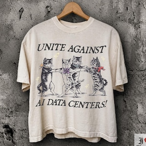 Op de afbeelding: Een ivoorkleurig T-shirt met korte mouwen en een afbeelding van vier katten die elkaar in een cirkel bij de pootjes vasthouden. De tekst erboven luidt UNITE AGAINST en eronder AI DATA CENTERS! De katten zijn zwart-wit getekend met kleine strikjes.