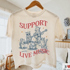 以下が含まれることがあります： アイボリーの半袖Tシャツ。アライグマ、キツネ、リス、カエル、ウサギが楽器を演奏しているグラフィックがプリントされています。イラストの上下には赤色で「SUPPORT LIVE MUSIC」という文字がデザインされています。