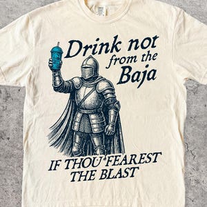 Peut inclure: Un t-shirt ivoire avec un motif bleu foncé représentant un chevalier en armure tenant une tasse bleue. Le texte indique : Drink not from the Baja IF THOU FEAREST THE BLAST. Le motif est centré sur la poitrine du vêtement à manches courtes.