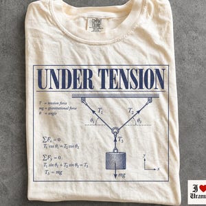Puede incluir: Una camiseta de color marfil con un estampado gráfico azul oscuro de un diagrama de física con la etiqueta UNDER TENSION. El diseño incluye un peso suspendido por dos cables, ecuaciones matemáticas de fuerza de tensión, gravedad y ángulos, además de un sistema de coordenadas.