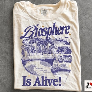 Könnte beinhalten: Ein elfenbeinfarbenes T-Shirt mit einem violetten Grafikdruck mit dem Text "Biosphere Is Alive!" und "Where air, water, and land meet." Das Design zeigt einen Querschnitt der Erde mit Bäumen, Bergen und verschiedenen geologischen Schichten.