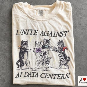 Pode incluir: Uma t-shirt cor de marfim com um gráfico de quatro gatos de mãos dadas num círculo. O texto diz UNITE AGAINST AI DATA CENTERS! Os gatos estão ilustrados a preto e branco com pequenos laços amarelos, roxos, azuis e vermelhos. Gola redonda.