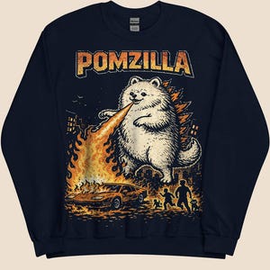 May include: Navy blue sweatshirt featuring a distressed graphic of a Pomeranian dog breathing fire, with the text "POMZILLA". The illustration includes a burning car, city skyline, and fleeing figures.