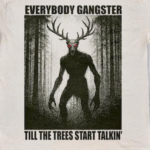 Op de afbeelding: Crèmekleurig T-shirt met een bosafbeelding met een silhouet met geweien en rode ogen. De tekst "EVERYBODY GANGSTER" en "TILL THE TREES START TALKIN'" staan boven en onder de afbeelding.
