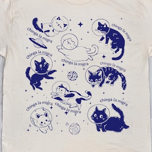 May include: Cream-colored t-shirt featuring a navy blue illustration of cats in space helmets, with the phrase "chinga la migra" repeated around the cats. The shirt has a crew neck and short sleeves. The text "NATURAL COLOR" is at the bottom.