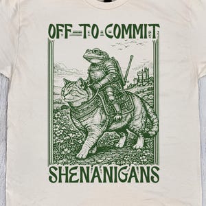 以下が含まれることがあります： クリーム色のTシャツ。緑色のイラストで、鎧を着たカエルが猫に乗っており、「OFF TO COMMIT SHENANIGANS」の文字が書かれています。このシャツは「NATURAL COLOR」と表示されています。