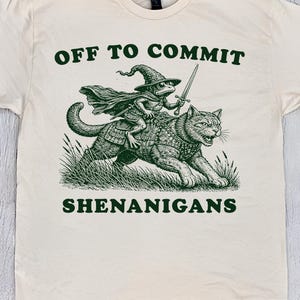 May include: Cream-colored t-shirt with a green graphic of a frog in a wizard hat riding a cat, holding a sword. Text reads "OFF TO COMMIT SHENANIGANS". The shirt is a natural color.