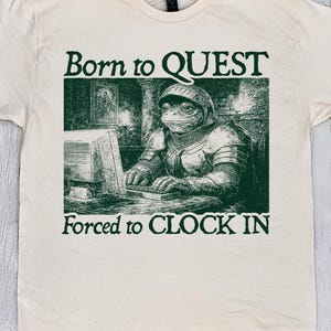 May include: A cream-coloured t-shirt featuring a green graphic of a frog in armour using a computer, with the text "Born to QUEST" and "Forced to CLOCK IN". The shirt is labelled "NATURAL COLOR".