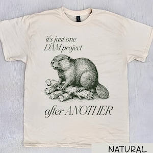 May include: Cream-colored t-shirt featuring a detailed illustration of a beaver sitting on a log. The text reads "it's just one DAM project after ANOTHER." The shirt is labeled "NATURAL COLOR."