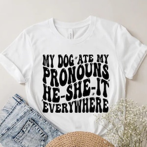 以下が含まれることがあります： 白いTシャツに黒い文字で「私の犬は私の代名詞を彼-彼女-それをどこでも食べた」と書かれています。