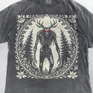 Pode incluir: Camiseta cor pimenta com uma figura esquelética com chifres e olhos vermelhos, emoldurada por um design de floresta e flores. O texto "I LOVE URANUSEVN" está na parte inferior.