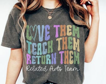 May include: Dark gray t-shirt with the words "LOVE THEM TEACH THEM RETURN THEM" in colorful, retro-style lettering. Below the text is "Related Arts Team" in a script font. The shirt has a vintage, worn look.