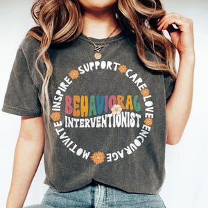 May include: Dark gray t-shirt with the words "Behavioral Interventionist" in a colorful, retro font. The words "Inspire, Support, Care, Love, Encourage, Motivate" are in a circle around the main text, with flower accents.