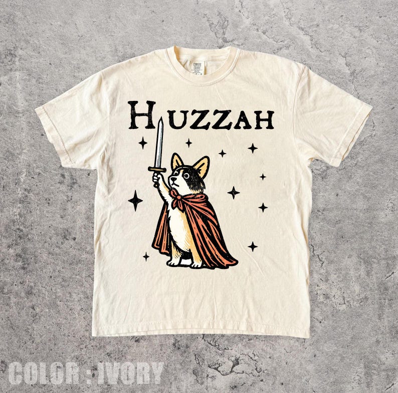K&ouml;nnte beinhalten: Elfenbeinfarbenes T-Shirt mit einem Cartoon-Corgi, der einen Umhang tr&auml;gt und ein Schwert h&auml;lt. Das Wort "Huzzah" ist &uuml;ber dem Corgi aufgedruckt, mit Sternen. Das T-Shirt ist aus weichem Material.