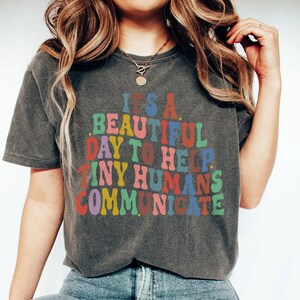 May include: A grey t-shirt with the text "It's a beautiful day to help tiny humans communicate" in colourful letters.