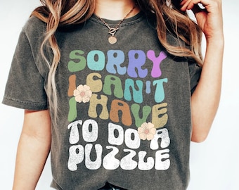 May include: A dark gray t-shirt with a retro-style graphic that reads "Sorry I can't I have to do a puzzle". The graphic is in rainbow colors with white flowers.
