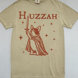 K&ouml;nnte beinhalten: Khakifarbenes T-Shirt mit roter Grafik eines Corgis mit Umhang und Schwert, dar&uuml;ber das Wort "Huzzah". Das Design enth&auml;lt Sterne. Ein l&auml;ssiges Kleidungsst&uuml;ck.