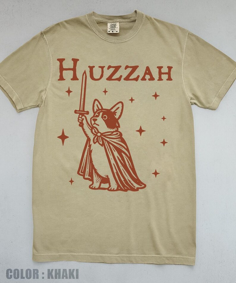 K&ouml;nnte beinhalten: Khakifarbenes T-Shirt mit roter Grafik eines Corgis mit Umhang und Schwert, dar&uuml;ber das Wort "Huzzah". Das Design enth&auml;lt Sterne. Ein l&auml;ssiges Kleidungsst&uuml;ck.