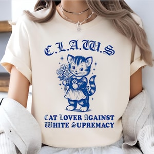 May include: Cream-colored t-shirt with a blue graphic of a cat holding flowers. The text above the cat reads "C.L.A.T.U.S." and below it says "CAT LOVER AGAINST WHITE SUPREMACY."