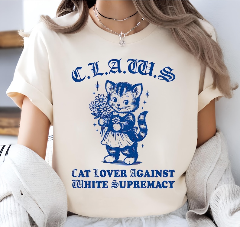 May include: Cream-colored t-shirt with a blue graphic of a cat holding flowers. The text above the cat reads "C.L.A.T.U.S." and below it says "CAT LOVER AGAINST WHITE SUPREMACY."
