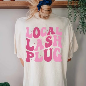 May include: A white t-shirt with a pink and light pink graphic that says "LOCAL LASH PLUG" in a retro font.