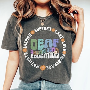 May include: A grey t-shirt with a circle design featuring the word "DEAF" in colourful letters. The circle also includes the words "Dream Team Education" and "Inspire, Support, Care, Love, Encourage, Motivate, Embrace".