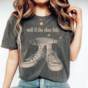 May include: Dark grey t-shirt with the phrase "well if the shoe fish." The shirt features a graphic of a fish wearing sneakers. The t-shirt is a casual, unisex garment.