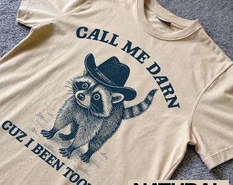 May include: Cream-colored t-shirt featuring a raccoon wearing a cowboy hat. The graphic includes the text "CALL ME DARN CUZ I BEEN TOOTIN'" in navy blue. The word "NATURAL" is printed at the bottom.