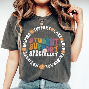 Può includere: T-shirt grigio scuro con la scritta "Student Support Specialist" in un font retrò colorato. Il design include una ghirlanda floreale con parole come "Inspire", "Support" e "Encourage". Un capo casual per tutti i giorni.