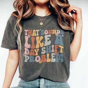 Könnte beinhalten: Ein dunkelgraues T-Shirt mit einer Retro-Grafik, auf der "That sounds like a day shift problem" steht. Die Grafik ist in bunten, abgerundeten Buchstaben mit Blumen.