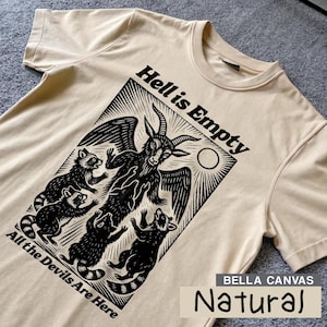 Puede incluir: Una camiseta Bella Canvas de color natural con un gráfico negro. El gráfico incluye una figura parecida a una cabra con alas, rodeada de mapaches, con el texto "Hell is Empty" y "All the Devils Are Here".