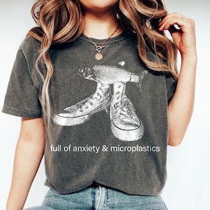 May include: Dark gray t-shirt with a white graphic of a fish and sneakers. The text "full of anxiety & microplastics" is printed below the graphic. The shirt is worn by a person.