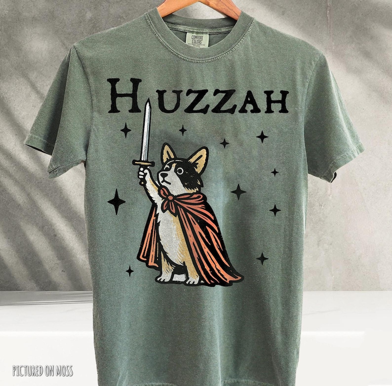 K&ouml;nnte beinhalten: Salbeigr&uuml;nes T-Shirt mit einem Cartoon-Corgi, der einen Umhang tr&auml;gt und ein Schwert h&auml;lt, mit dem Wort "Huzzah" dar&uuml;ber. Sterne erg&auml;nzen das Design. Das T-Shirt h&auml;ngt an einem Holzkleiderb&uuml;gel.