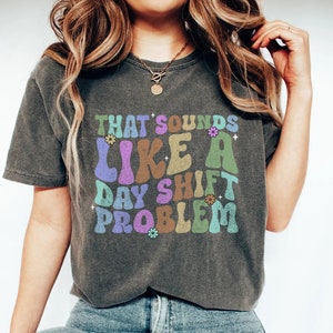 May include: A dark grey t-shirt with a retro-style graphic that reads "That sounds like a day shift problem". The graphic is in various colours, including purple, green, blue, and orange, with small flower accents.