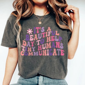 Puede incluir: Una camiseta gris con un gráfico rosa, morado y azul que dice "Es un hermoso día para ayudar a los pequeños humanos a comunicarse".