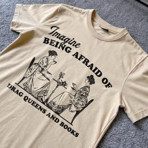 Op de afbeelding: Crèmekleurig T-shirt met een zwarte afbeelding. De afbeelding toont twee vrouwen aan een tafel die thee drinken, met de tekst "Imagine Being Afraid of Drag Queens and Books."