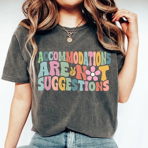 Puede incluir: Una camiseta gris oscuro con el texto "Accommodations Are Not Suggestions" en letras coloridas de estilo retro. El texto incluye un signo de paz, una pelota de baloncesto y una flor.